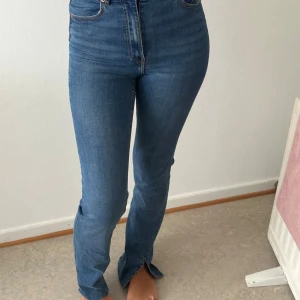 Blå high waist jeans från Gina Tricot - Säljer ett par klassiska blå jeans från Gina Tricot med hög midja och raka ben. Jeansen har snygg slitning, fem fickor och en liten slits nertill vid benslut. Perfekt passform och stretchigt material för en bekväm känsla.