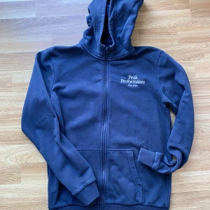 Mörkblå hoodie Peak Performance - Mörkblå hoodie från Peak Performance med dragkedja och huva. Broderad logga på bröstet och två stora fickor framtill. Skön och mjuk insida, perfekt för kyliga dagar. Klassisk passform och ribbade muddar vid ärmslut och nederkant.