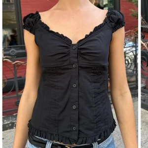 Svart blus med volang och knappar - Supersöt svart blus med volangdetaljer längs urringning och nederkant. Knappar framtill och korta puffärmar ger en romantisk vibe. Figurnära passform som framhäver midjan, perfekt till jeans eller kjol.