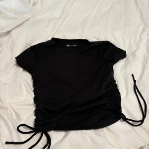 Svart ribbad topp med snörning - Svart ribbad topp från Zara med korta ärmar och cool snörningsdetalj på båda sidorna som gör att du kan justera längden. Perfekt till jeans eller kjol för en trendig look. Enkel att matcha och superbekväm.