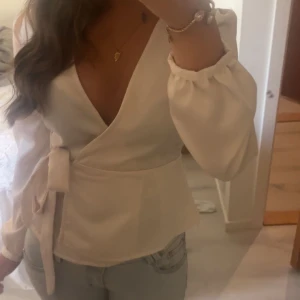 Beige/champagne/rosa omlottblus med knytband - Säljer en stilren omlottblus med v-ringning och långa puffärmar. Blusen har ett brett knytband i midjan som ger en snygg siluett. Perfekt att matcha med jeans eller kjol för en trendig look. Satin, märket är glamourous! Storlek xs-s