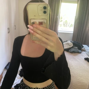 Svart croppad topp från Bershka - Svart långärmad croppad topp från Bershka med fyrkantig urringning och lätt ribbad struktur. Ärmsluten har vågig kant för en extra detalj. Perfekt att styla med höga jeans eller kjol för en trendig look. 