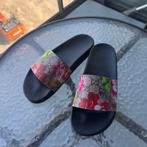 Sandaler med blommigt mönster - Svarta sandaler från  med bred rem i canvas, dekorerad med färgglada blommor och klassiskt GG-mönster. Perfekta för sommaren med sin platta sula och trendiga design. Snyggt statement-piece för dig som gillar lyxiga detaljer.