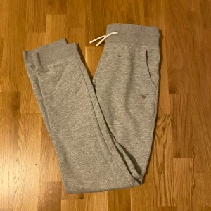 Grå mjukisbyxor från Gant - Snygga grå mjukisbyxor från Gant med dragsko i midjan och ribbade muddar vid benslut. Byxorna har fickor på sidorna och en liten broderad Gant-logga på låret. Perfekta för chill eller träning.