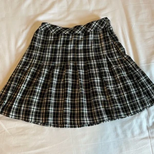 Svartvit rutig kortkjol med knäppning - Supersnygg svartvit rutig kortkjol med gul detaljtråd och klassiskt veckad design. Kjolen har en silvrig knapp i midjan och är perfekt för en cool och trendig look. Passar grymt till sneakers eller boots.