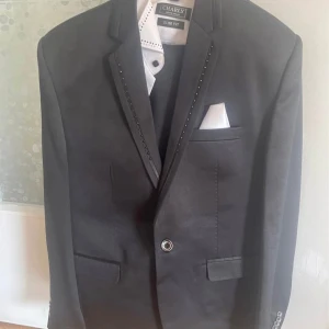 Svart kavaj från Ceniea Italy slim fit - Snygg svart kavaj från Ceniea Italy i slim fit-modell. Kavajen har dekorativa nitar längs slaget, klassiska fickor och vit näsduk i bröstfickan. Perfekt för dig som vill ha en stilren och modern look.