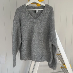 Grå stickad v-ringad tröja Divided - Mysig grå stickad tröja från Divided med djup v-ringning och breda ribbade muddar. Perfekt för lager-på-lager och chill dagar. Oversized fit som ger en avslappnad vibe. Passar dig som gillar enkel och stilren look.