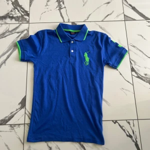 Blå pikétröja från Ralph Lauren S - Snygg blå pikétröja från Ralph Lauren med gröna detaljer på krage och ärmslut. Ikonisk grön polospelare broderad på bröstet. Klassisk pikékrage, korta ärmar och knäppning framtill. Perfekt för en sportig och stilren look.