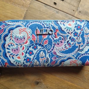 LiLiÓ plånbok - En plånbok från LiLiÓ med fina mönster i blått, rosa och vitt. Plånboken har ett fack med dragkedja och flera kortfack inuti. Varan har inga eller små tecken på användning. Storlek längd 18cm bredd 10cm Material : polyester