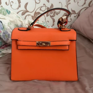 Orange handväska med gulddetaljer - Snygg orange handväska med stilren, boxig form och guldfärgat spänne framtill. Väskan har ett kort handtag med svarta kanter och en dekorativ scarf knuten vid fästet. Perfekt för dig som vill sticka ut med färg och elegans.