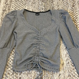 Rutig svartvit topp från Gina Tricot - Supersnygg svartvit rutig topp från Gina Tricot i croppad modell. Toppen har långa puffärmar, v-ringning och snörning framtill som ger en rynkad effekt. Storlek XS