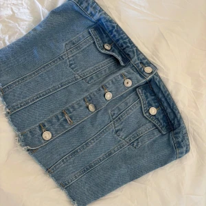 Blå jeanstopp med knappar - Snygg kort jeans topp i ljusblå denim med fransig nederkant, knappar framtill och elastisk smock baktill för extra komfort. Passar jättefint till ett par jeans eller kjol. Passar både S-M
