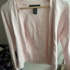 Ljusrosa ribbad kofta från Banana Republic - Supersöt ljusrosa kofta från Banana Republic i ribbad bomull. Klassisk modell med knappar framtill och långa ärmar. Perfekt att slänga över en topp när det är lite kyligt ute. Enkel och stilren design som funkar till det mesta.