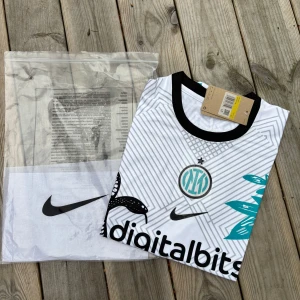Inter Milan Nike vit bortatröja 2022/23 - Inter Milan bortatröja från Nike i vitt med svarta och turkosa detaljer. Tröjan har korta ärmar, svart krage och är prydd med ormmotiv, grafiska linjer och klubbmärke på bröstet. Materialet är lätt och andas, perfekt för fotboll eller supporterliv.