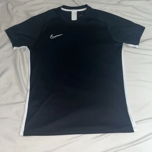 Svart Nike Dri-Fit t-shirt herr M-L - Svart Nike Dri-Fit t-shirt med vita detaljer längs sidorna och axlarna. Klassisk sportig look med korta ärmar och rund hals. Perfekt för träning med lätt och snabbtorkande material. Swoosh-logga på bröstet för extra stil.