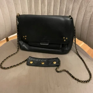 Svart axelväska från Jerome Dreyfuss - Stilren svart axelväska i skinn från Jerome Dreyfuss med guldfärgade nitar och kedjerem. Väskan har lock med tryckknapp, ytterfack med dragkedja och en avtagbar axelrem med snygga detaljer. Perfekt för alla tillfällen🥰