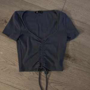 Blå croppad topp med snörning från ZARA - Snygg blå croppad topp från ZARA med korta ärmar och djup v-ringning. Toppen har ribbad struktur och snörningsdetalj. Storlek XXS