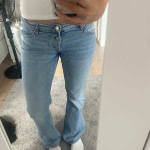 Ljusblå bootcut jeans med knappar - Säljer ett par ljusblå jeans med bootcut passform och låg midja. Jeansen har flera synliga knappar framtill och klassisk femficksdesign. Jeansen är ifrån Gina Young och är i storlek 170.