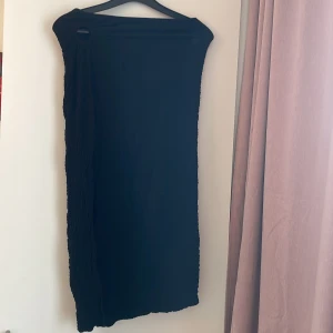 Svart offshoulder klänning med ringdetalj - Svart, axelbandslös klänning från NK Mode med snygg ringdetalj vid axeln. Klänningen har en skrynklig textur och rak passform som slutar vid knäna. Perfekt för dig som gillar minimalistisk och stilren look.