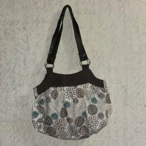 Blommig axelväska med bruna detaljer - Snygg axelväska med blommigt mönster i brunt, beige och grönt på vit botten. Väskan har bruna handtag i skinnimitation och brunt tyg upptill. Insidan är fodrad i glansigt brunt tyg och stängs med magnetknapp. Perfekt storlek för vardagens prylar.