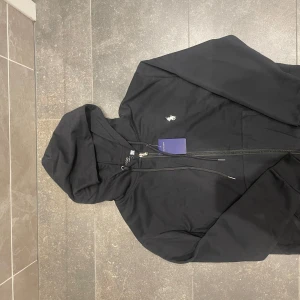 Svart hoodie från Polo Ralph Lauren - Svart hoodie från Polo Ralph Lauren med klassisk broderad logga på bröstet. Tröjan har huva med snörning, dragkedja framtill och känguruficka. Tillverkad i mjuk bomull för en skön och avslappnad stil. Perfekt för dig som gillar stilrena och tidlösa plagg.