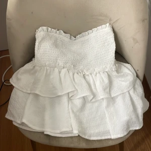 Vit volang topp med smock - Supersöt vit kort topp med smockad överdel och volanger i kjolen. Axelbandslös modell med rynkade detaljer upptill och dubbla lager volang nedtill. Perfekt för sommarens alla tillfällen och ger en fräsch och luftig känsla. ÄR EN TOPP INTE KJOL!!🩷