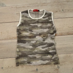 Camo mesh linnetopp från Koola Anna - Säljer en ärmlös mesh-linnetopp från Koola Anna i storlek S. Toppen har ett coolt camouflage-mönster i grått, vitt och beige med vita kanter runt hals och ärmhål. 