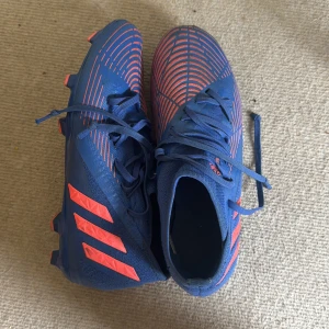 Blå och röda adidas fotbollsskor - Säljer ett par adidas fotbollsskor i blått med röda detaljer och mönster. Skorna har snörning, platt sula med dobbar och är tillverkade i syntetmaterial. Perfekta för dig som vill sticka ut på planen med snygg design och bra grepp.