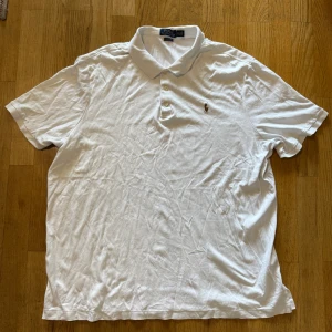 Vit pikétröja från Polo Ralph Lauren XXL - Klassisk vit pikétröja från Polo Ralph Lauren i storlek XXL. Tröjan har korta ärmar, krage med tre knappar och den ikoniska broderade polospelaren på bröstet. Classic fit-modell i mjuk bomull, perfekt för en clean och stilren look.