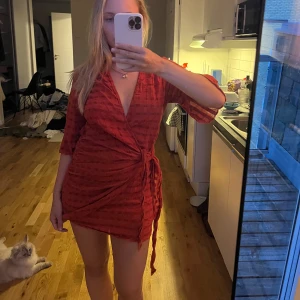 Röd omlottklänning med knyt - Säljer en kort röd omlottklänning med knyt i midjan och trekvartsärm. Klänningen har ett diskret rutigt mönster och v-ringad hals. Perfekt för dig som gillar en feminin och trendig look. Materialet känns lätt och skönt mot huden.