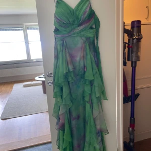 Grön långklänning med volanger - Säljer en magisk långklänning i grönt med lila och ljusa inslag. Klänningen har tunna axelband, omlott-detaljer över bysten och vackra volanger längs kjolen. Materialet är lätt och flowy, perfekt för sommarkvällar. Sidoskärningar ger en extra snygg touch.