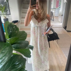 Blommig långklänning med volang - En långklänning med blommigt mönster i ljusa pastellfärger. Klänningen har tunna axelband, volangdetaljer vid bysten och en öppen rygg. 