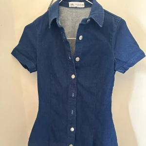 Mörkblå jeansklänning från Zara - Supersnygg mörkblå jeansklänning från Zara med korta ärmar, klassisk krage och knappar hela vägen framtill. Figurnära passform som ger en clean och cool look. Perfekt för dig som gillar denim och vill ha något enkelt men stilrent.