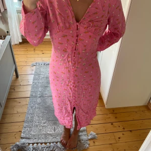 Rosa blommig långklänning med knappar - Supersöt rosa långklänning med små blommor i vitt och orange. Klänningen har långa ärmar, v-ringning och knappar hela vägen framtill. Den är för stor för mig så den ska sitta som den gör på första bilden egentlige!💞