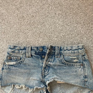 Blå jeansshorts med fransig kant - Snygga blå jeansshorts med låg midja och slitna detaljer. Shortsen har fransig kant nertill, klassiska fickor fram och bak samt dubbla knappar i midjan. Perfekta för varma dagar och ger en cool, avslappnad vibe.