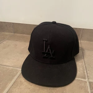 Svart LA-keps New Era 59FIFTY - Snygg svart keps från New Era 59FIFTY med LA-logga framtill i svart. Klassisk modell med böjd skärm och broderade detaljer. Tillverkad i 100% polyester och en del av Cooperstown Collection. Perfekt för dig som gillar streetstyle.