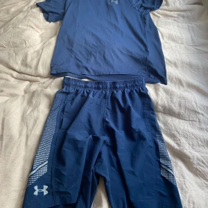 Blåt Under Armour set  - Blå t-shirt och shorts från Under Armour i loose fit, perfekt för träning. Klassisk kortärmad modell med diskret logga på bröstet. Tillverkad i lätt och funktionell polyester som andas och håller dig sval under intensiva pass.