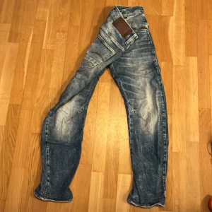 Blå jeans från G-Star RAW - Säljer ett par blå jeans från G-Star RAW med snyggt slitna detaljer och markerade sömmar. Jeansen har fem fickor, normal passform och coola sömmar över knäna som ger en unik look. Perfekta för dig som gillar streetstyle och vill sticka ut.