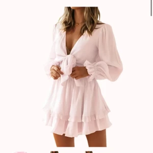 Ljusrosa klänning med volang från design by Amor - Supersöt ljusrosa kortklänning med och knyt framtill. Klänningen har långa ärmar med lätt puff och volangdetaljer i nederkant. Djup v-ringning och flowy passform ger en romantisk vibe. Perfekt för vår och sommar! Orginalpris 299kr