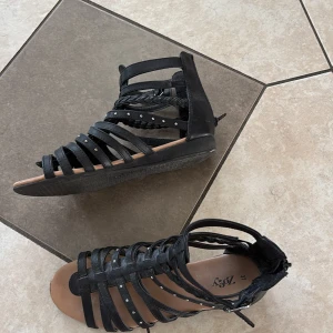 Svarta sandaler från Zoey, stl 37 - Snygga svarta sandaler från Zoey med flätade och släta remmar, dekorerade med små silverfärgade nitar. Skorna har öppen tå, platt sula och dragkedja bak för enkel påtagning. Perfekta för varma dagar när du vill ha en cool och trendig look.