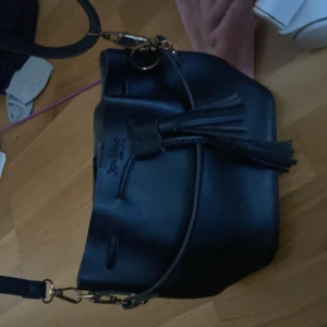 Svart axelväska från Joe Mac - Snygg svart axelväska från Joe Mac med stilren design och dekorativ tofs framtill. Väskan är gjord i slätt skinn och har justerbar axelrem i matchande färg. Perfekt storlek för att få plats med allt du behöver och enkel att matcha med olika outfits.