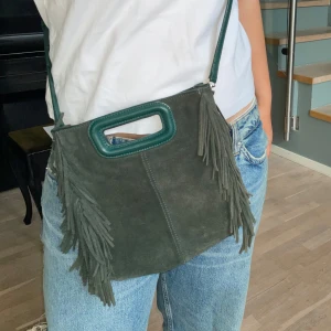 MAJE "Fringed M Bag" i mörkgrön mocka - Snygg väska från Maje i mörkgrön mocka med fransar. Modellen heter Fringed M Bag in suede och är en klassiker. Nypris cirka 3 800 kr.  Väskan är i fint skick överlag, men axelremmen har slitits(se bild 5) och ett av metallfästena har lossnat (se bild 4). Skadan går att laga hos skomakare, uppskattad kostnad ca 200–300 kr. Därför säljs den med lägre pris.  Specifikationer: Äkta mocka Handtag och avtagbar axelrem Fransdetaljer på båda sidor Magnetstängning.    Kommer i original påse.
