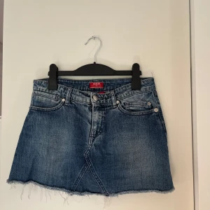 Blå jeanskjol mini - Snygg blå jeanskjol från Rager med rå, fransig nederkant och klassiska fem fickor. Baksidan har coola detaljer med strass på bakfickorna. Minikjol. 