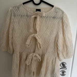 Beige blus med knyt från Gina Tricot - Supersöt beige blus från Gina Tricot med tre knyt framtill och puffiga halvlånga ärmar. Tunt, strukturerat tyg med vågigt mönster. Superfin till både sommar, höst och vinter och till både vardagar och festtillfällen💐Har aldrig använts.