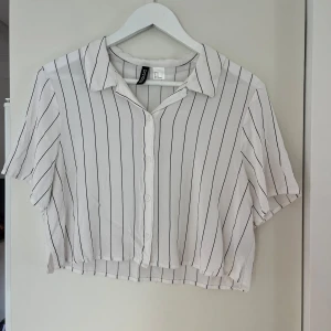 Vit randig croppad skjorta H&M Divided - Snygg croppad skjorta från H&M Divided i vitt med svarta vertikala ränder. Skjortan har korta ärmar, klassisk krage och knäppning framtill. Nästan aldrig använd. Väldigt snygg till skjorts och långkjol.