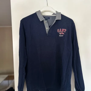 Marinblå pikétröja från GANT - Snygg marinblå långärmad pikétröja från GANT med grå krage och knappar framtill. Broderad GANT-logga med amerikansk flagga på bröstet. Perfekt för en preppy look och tillverkad i mjuk bomull. Nypris 1299kr