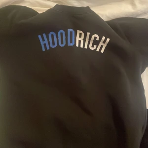 Svart Hoodrich sweatshirt med tryck - Svart sweatshirt från Hoodrich med HOODRICH-tryck i blått och vitt på ryggen. Tröjan har rund hals, långa ärmar och är gjord i en mjuk bomulls- och polyestermix. Perfekt för dig som gillar streetwear och vill ha en clean look.