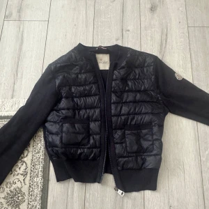 Svart quiltad cardigan jacka Moncler - Svart cardigan jacka från Moncler med quiltad front och två fickor. Jackan har långärmade stickade ärmar, dragkedja framtill och klassisk Moncler-logga på ena ärmen. Perfekt för dig som vill ha en stilren och exklusiv look. Dragkedjan fungerar inte.