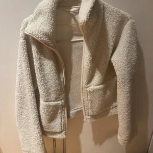 Beige teddyjacka från H&M, storlek S - Supermjuk och fluffig teddyjacka från H&M i beige. Jackan har dragkedja framtill, två stora fickor och en croppad passform. Perfekt för att hålla sig varm och ändå se chill ut. Långärmad och med hög krage för extra mysfaktor.