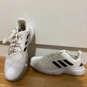 Vita Adidas sneakers med svarta ränder - Fräscha vita Adidas sneakers med klassiska svarta ränder på sidorna. Skorna har snörning och är tillverkade i mesh och syntet för en sportig look. Perfekta för dig som gillar stilrena och bekväma skor till vardagen eller träningen.